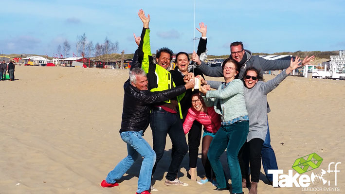 Een trots team dat hun Reuzevlieger net heeft opgelaten op het strand van Scheveningen. De vlieger staat in de lucht en ze hebben met zijn allen de vliegerhaspel vast.