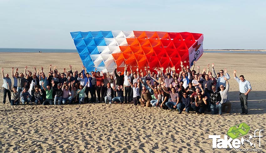 Een groepsfoto op het strand met in het midden een gigantische Megavlieger in 4 kleuren. De vlieger hebben de mensen net gebouwd tijdens een teambuilding workshop.